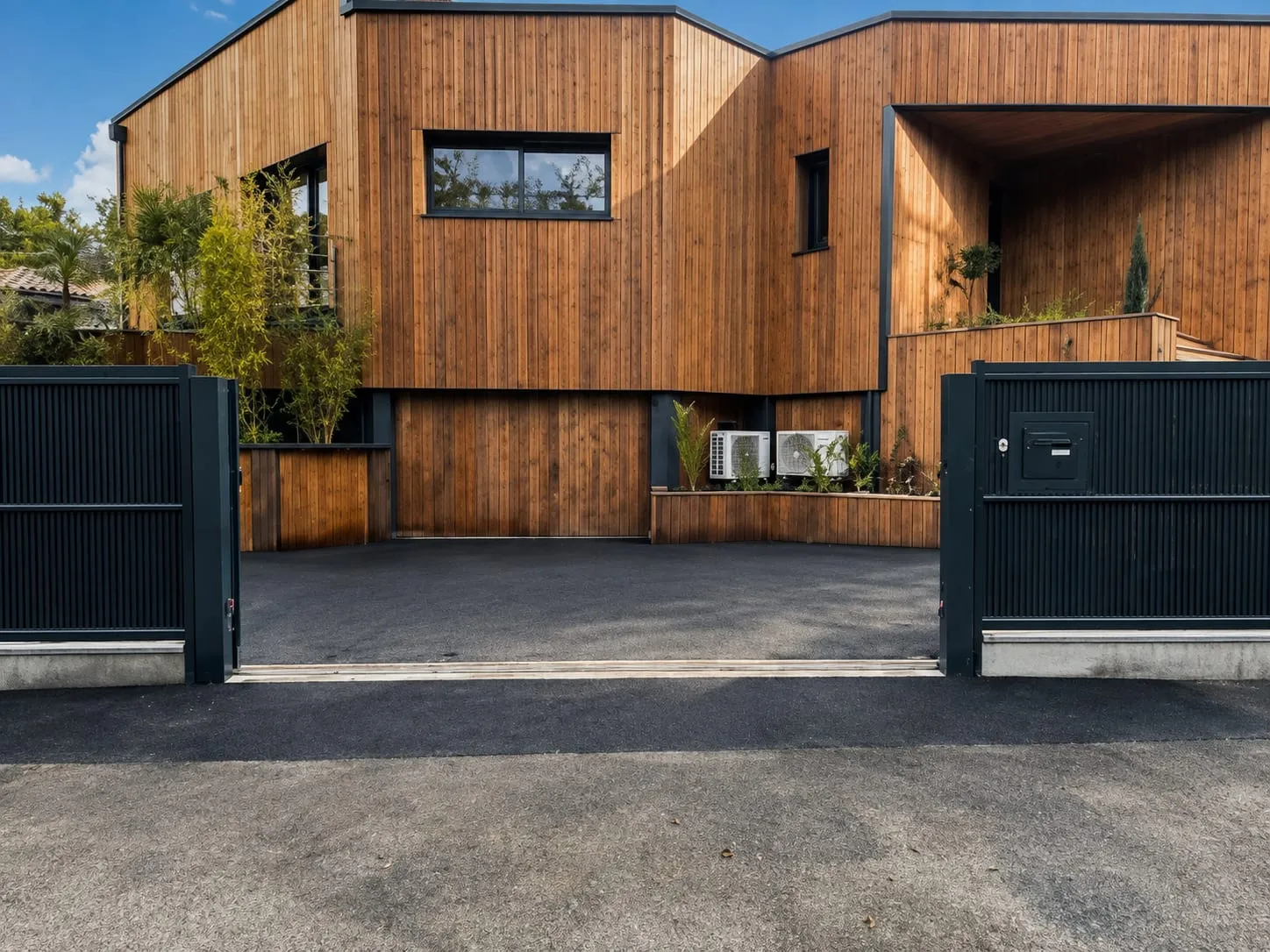 Cour en enrobé noir devant une maison au bardage bois et un portail métallique