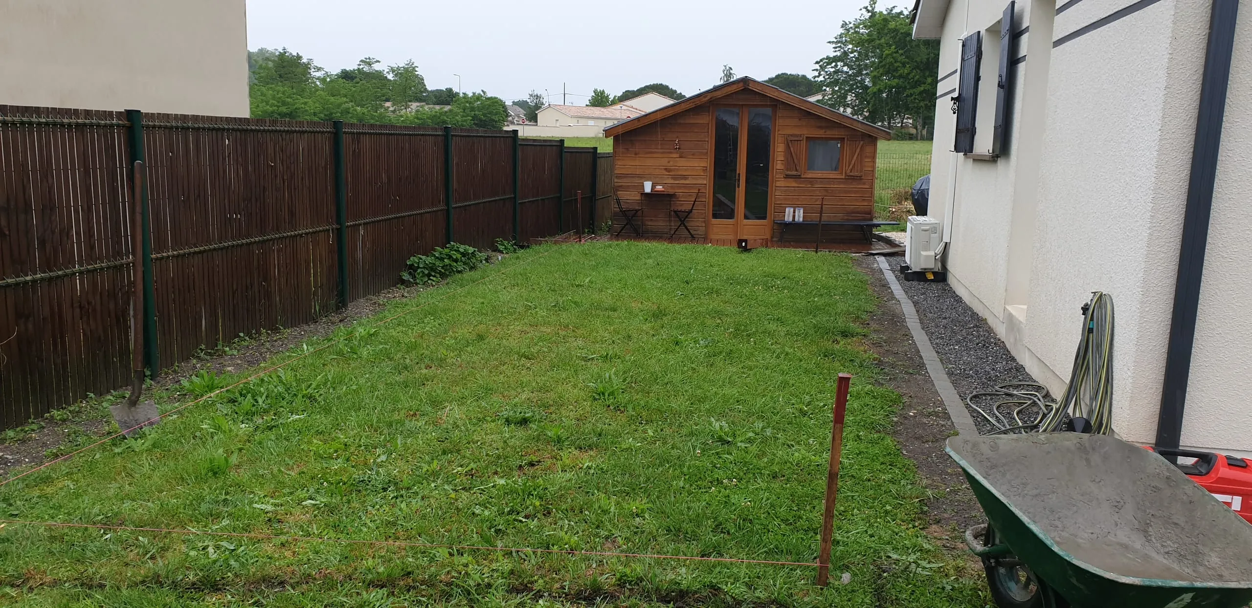 Jardin résidentiel clos par une clôture en lames de bois brun sur poteaux métalliques verts, avec abri de jardin en bois en fond de parcelle