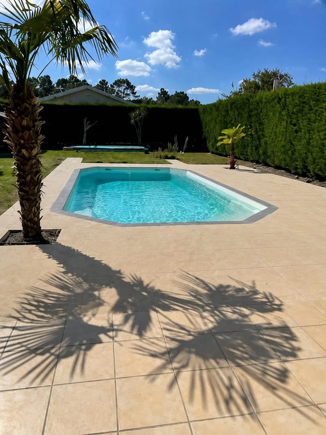 Terrasse carrelée beige autour d’une piscine dans un jardin bordé de haies